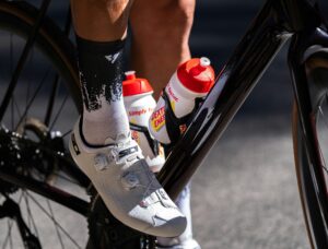 Cyclist's shoe on pedal with energy drink bottles attached to bike frame.